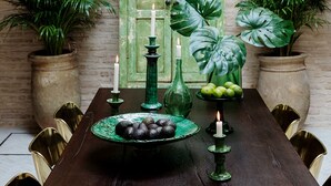 A dining table with a rustic wooden finish, surrounded by chairs, set against a brick wall with a green door.