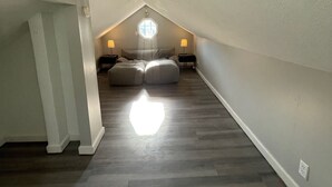 Une petite chambre mansardée, étroite et à pente, avec un puits de lumière, un plancher en bois et un lit agrémenté d’oreillers et d’une lampe.