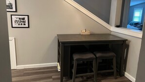 Un petit bureau en bois sombre, accompagné de deux tabourets assortis, est placé dans un coin d’une pièce aux murs gris et au plafond en pente.