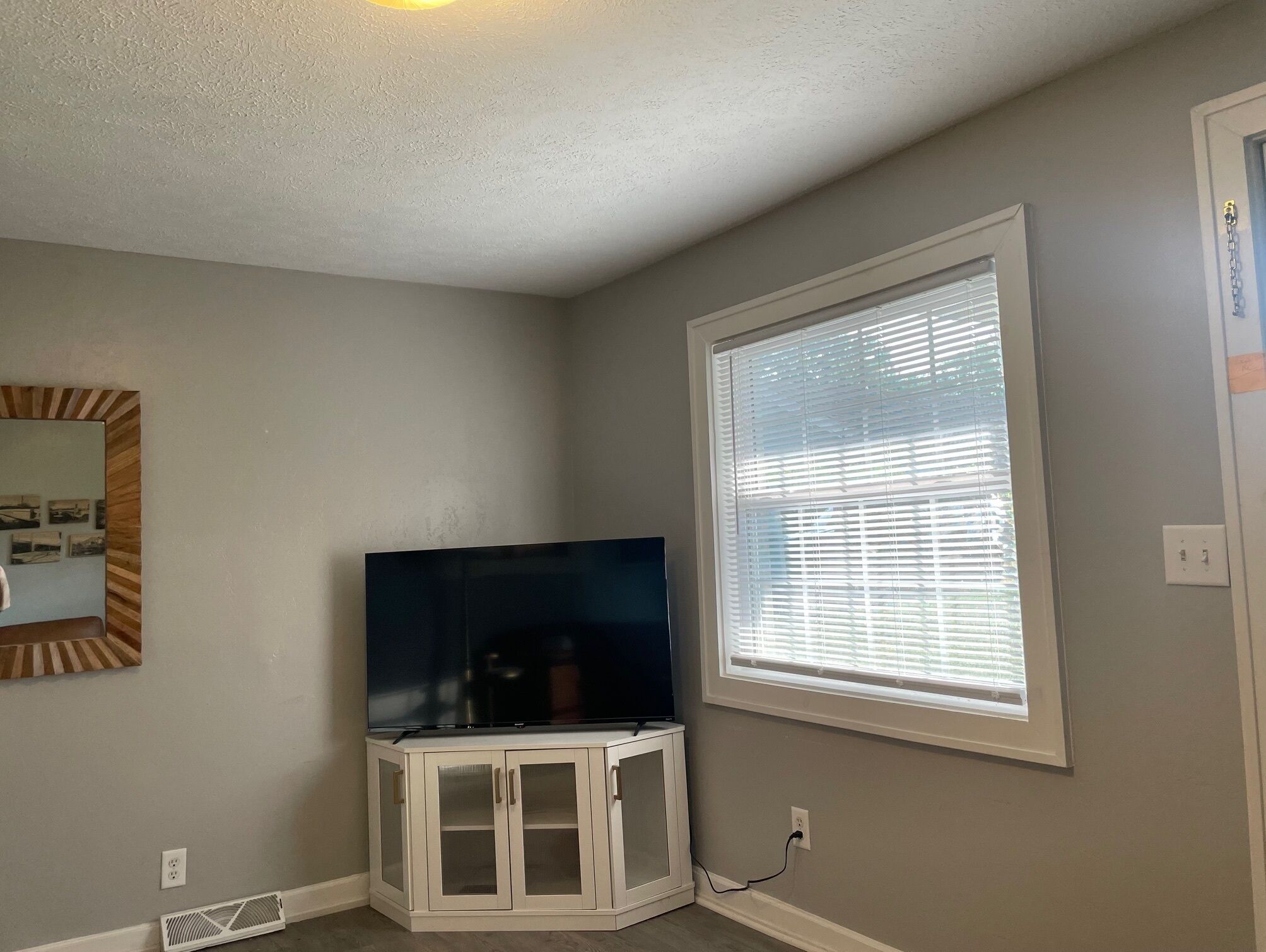 A room with a flat-screen TV on a white cabinet, a window with blinds, and a wall-mounted mirror.