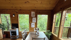 A wooden dining area with a table and chairs, surrounded by large windows with a view of greenery. -  Eureka Springs Riverview Oasis: A Serene Retreat Overlooking the White River  (Eureka Springs)