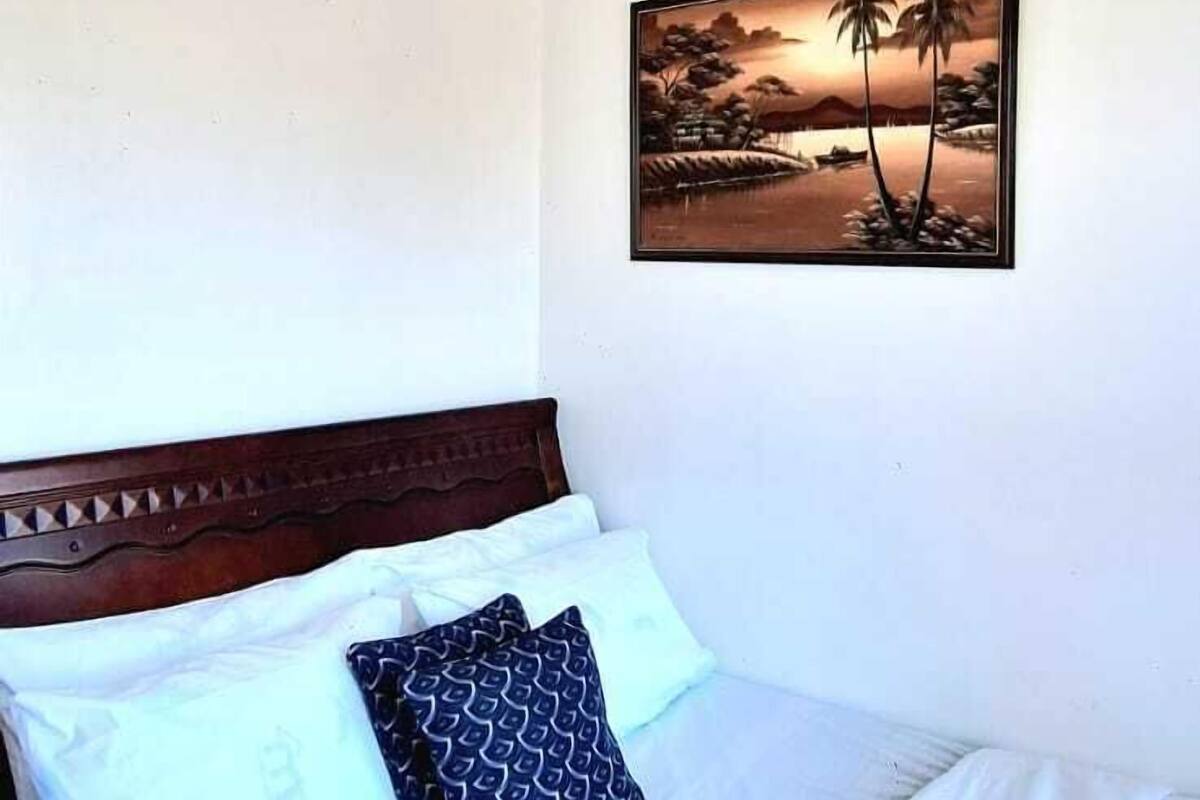 Un lit bien fait, avec du linge de lit blanc et des coussins à motifs, une tête de lit en bois et un tableau représentant un coucher de soleil, accroché au mur.