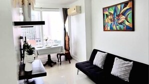 Un saló modern amb un sofà negre, un quadre a la paret i una zona de cuina amb finestra.