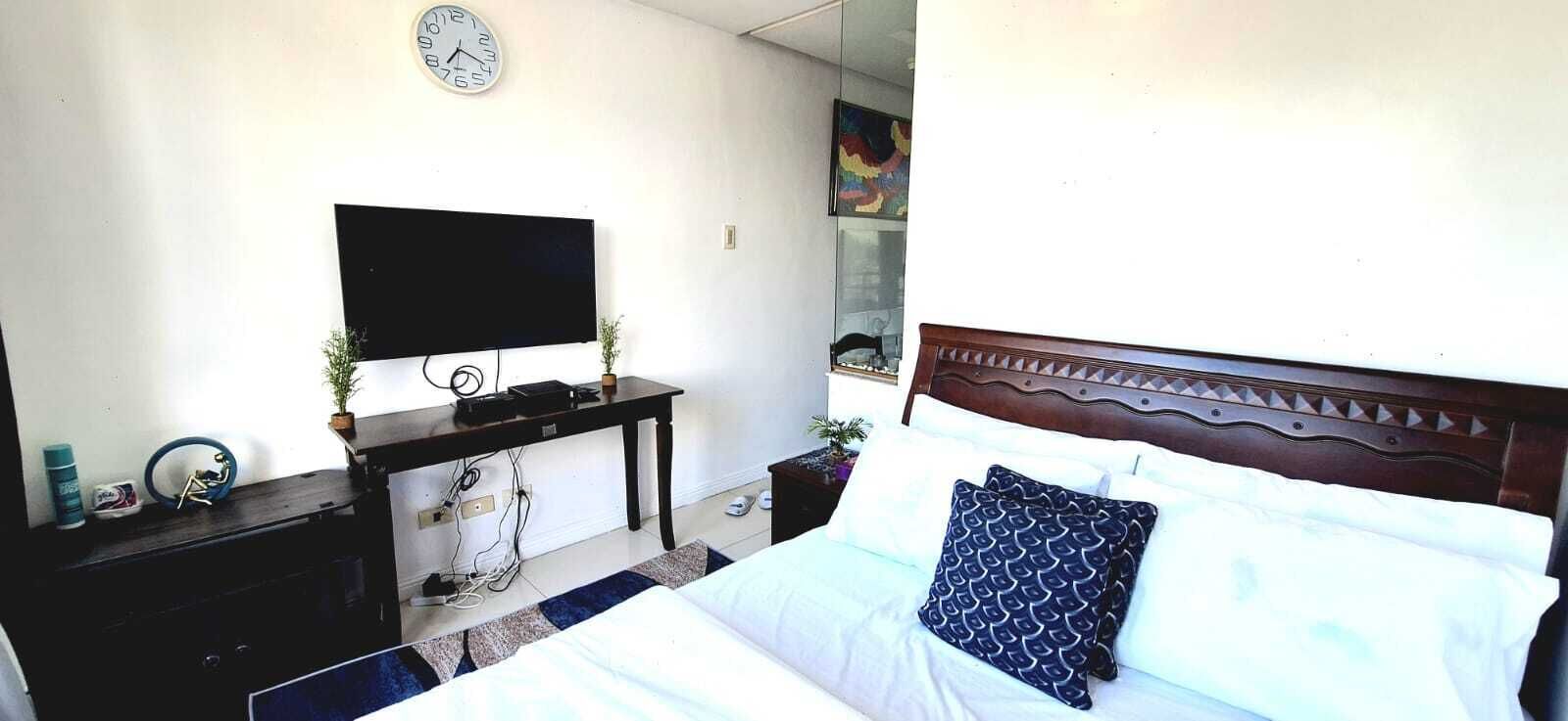 A hotel room with a bed, a TV on a stand, a desk with a computer, and a clock on the wall.