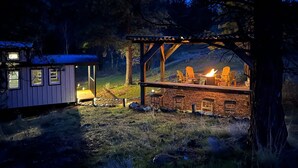 A rustic cabin with a small attached shed, a wooden deck with chairs and a fire pit, surrounded by trees and a mountain backdrop. - Sagebrush Caboose (Oliver)