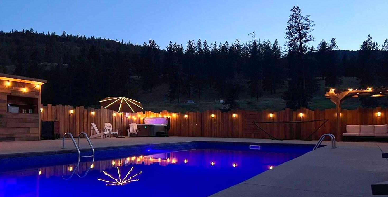 A swimming pool with a wooden deck, a barbecue area, and a wooden fence with string lights.