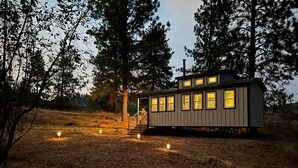 A small cabin with a porch and multiple windows, surrounded by trees and a forested area. - Sagebrush Caboose (Oliver)