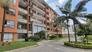 Un edificio residencial de varios pisos con balcones, entrada pavimentada y abundante vegetación, incluyendo palmeras.