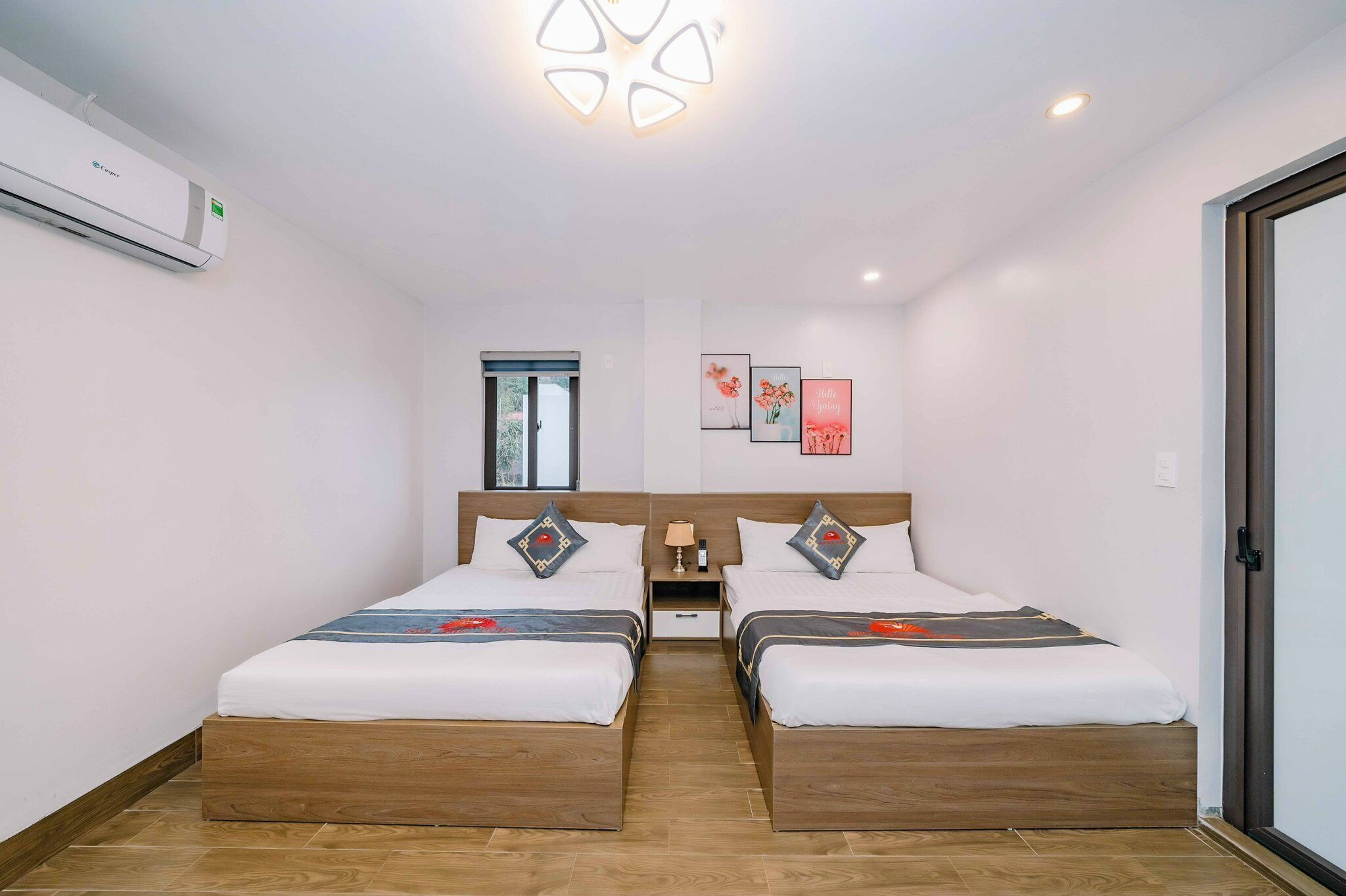 A hotel room with two single beds, a nightstand with a lamp, a window, and a wall-mounted air conditioner.