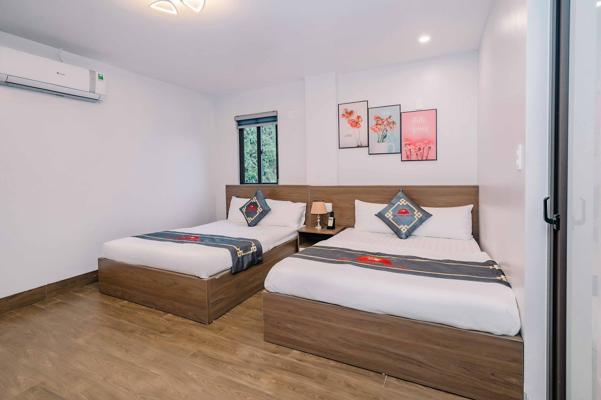 A hotel room with two beds, a window, wall art, and an air conditioning unit.