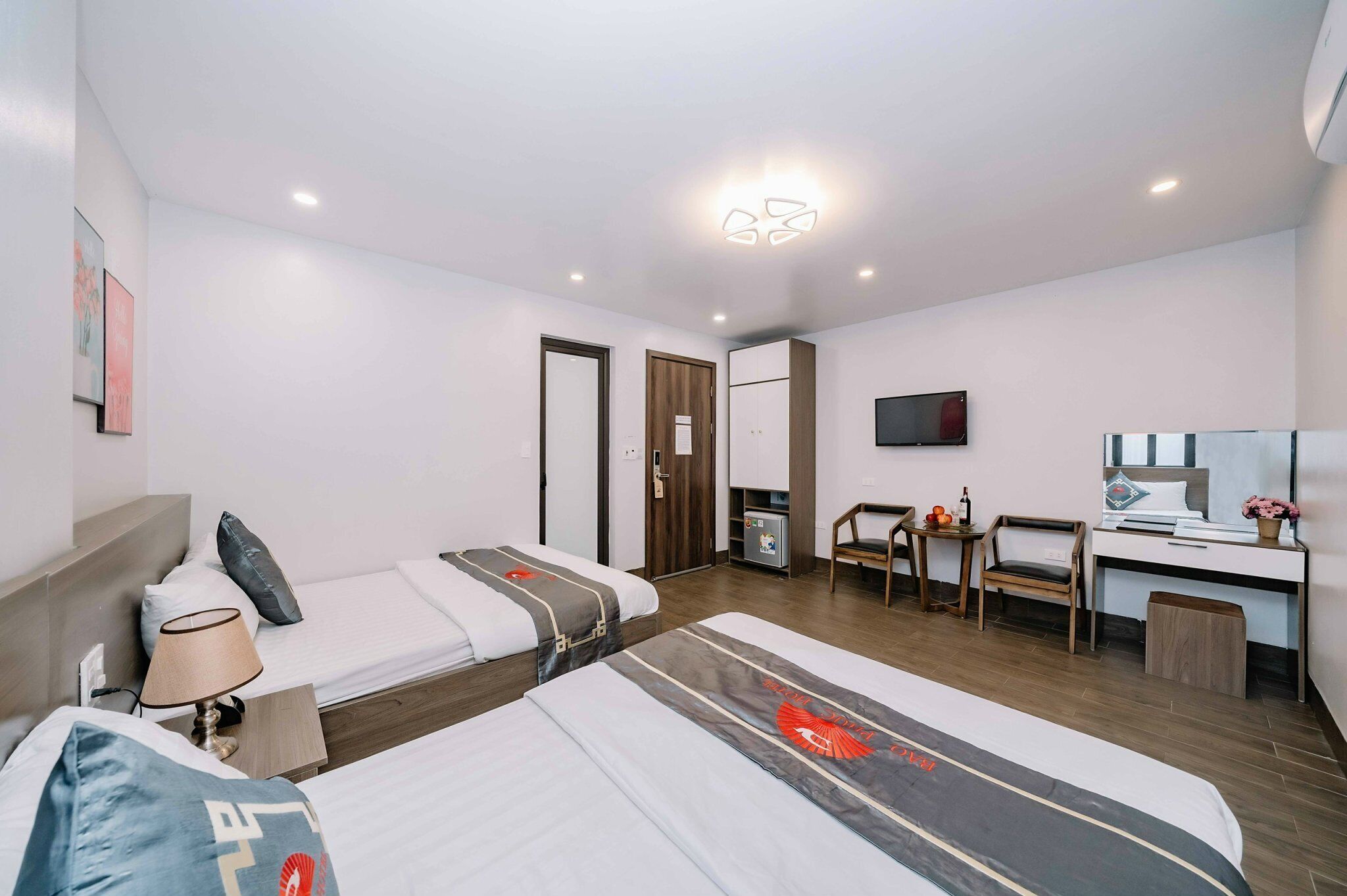 A modern hotel room with two single beds, a desk, a chair, a TV, and a wardrobe.
