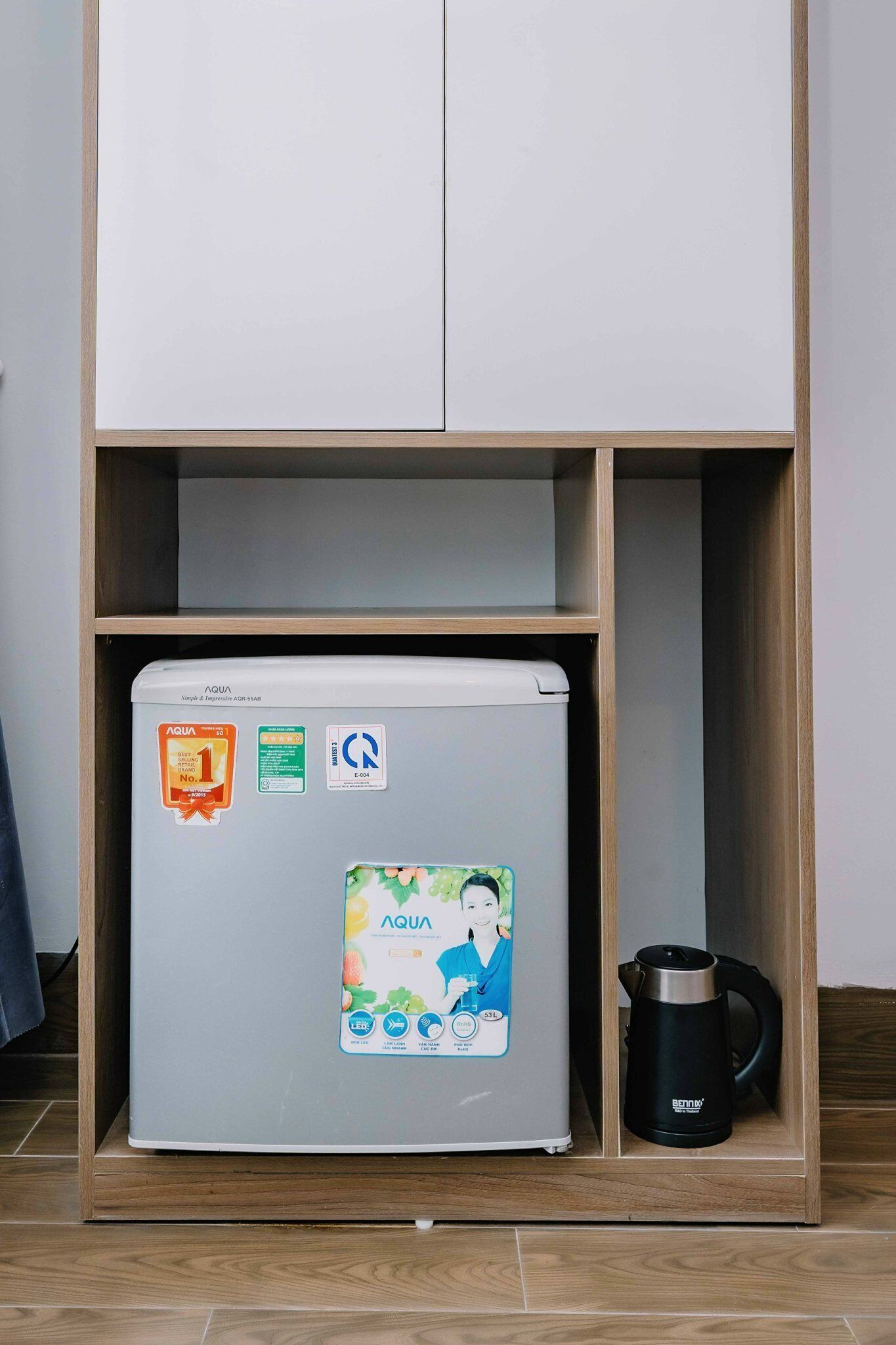 A compact refrigerator with a water dispenser, a kettle, and a cabinet above it.