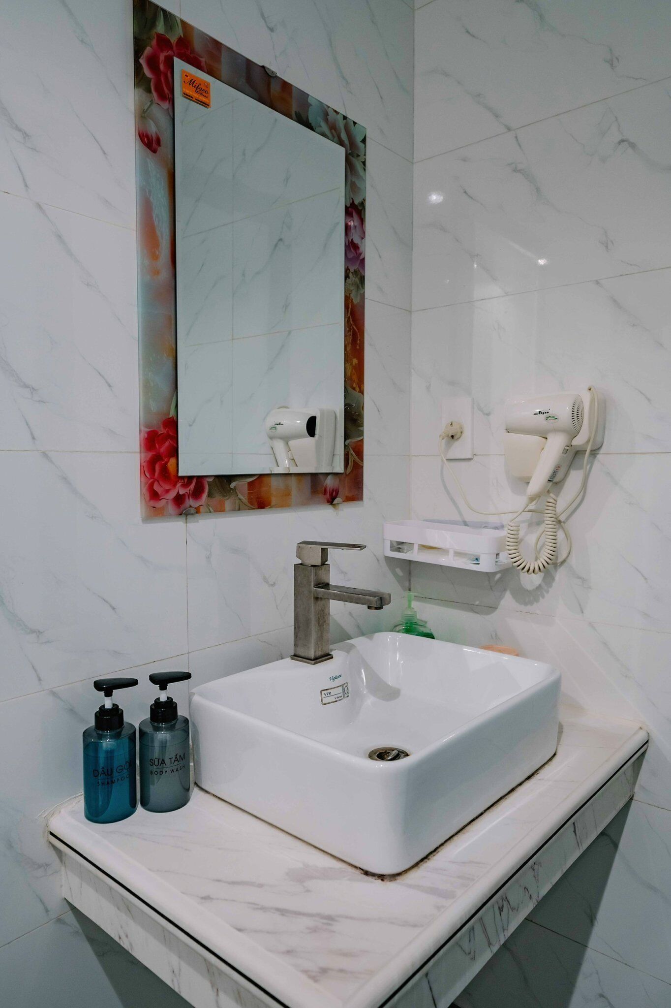 Un baño con lavamanos blanco, un espejo con marco floral, un secador de pelo y dos botellas de gel de ducha.