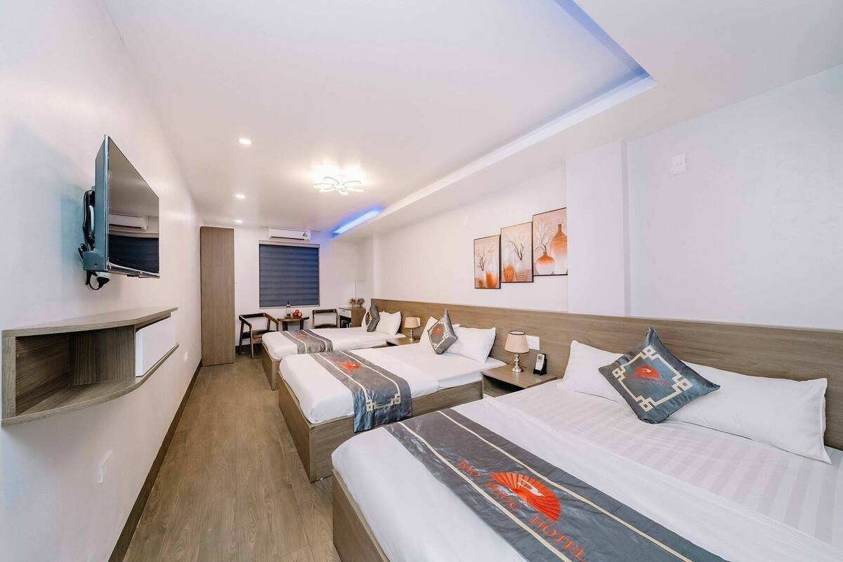 Ein Hotelzimmer mit drei Betten, einem an der Wand befestigten Fernseher, einem Schreibtisch und einem Sessel.