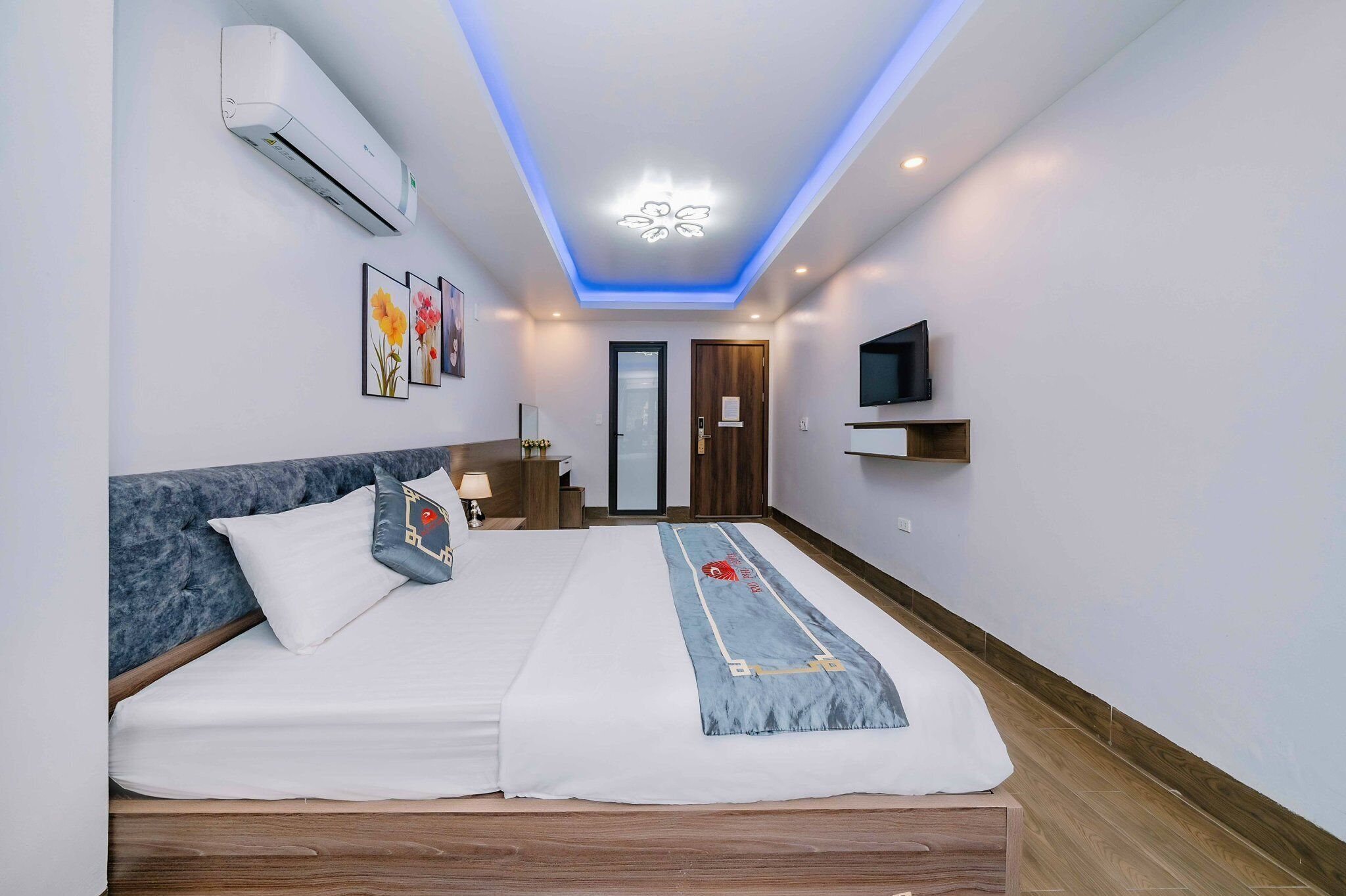 A modern hotel room with a bed, bedside table, lamp, air conditioning unit, artwork, and a television.