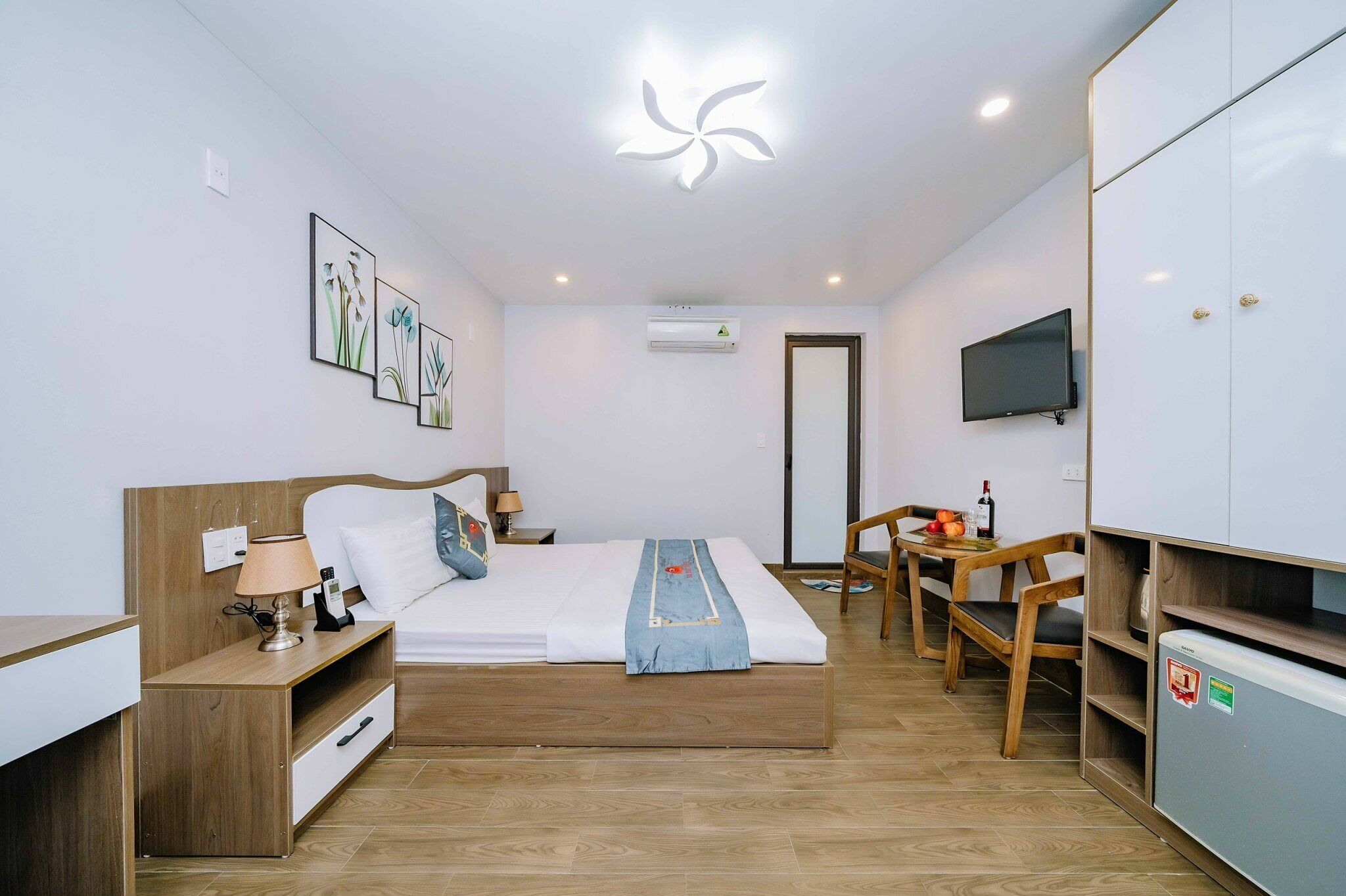 A modern hotel room with a wooden bed, a desk, a chair, a TV, and a mini-fridge.