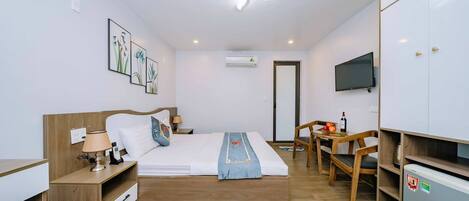 A modern hotel room with a wooden bed, a desk, a chair, a TV, and a mini-fridge.