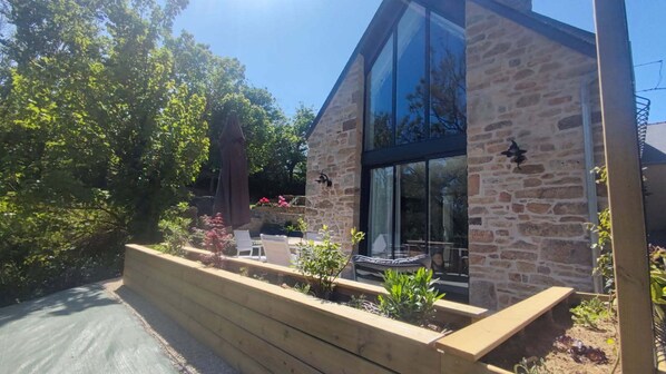 A modern building with stone walls and large glass windows, surrounded by a landscaped garden with a wooden bench and outdoor seating.