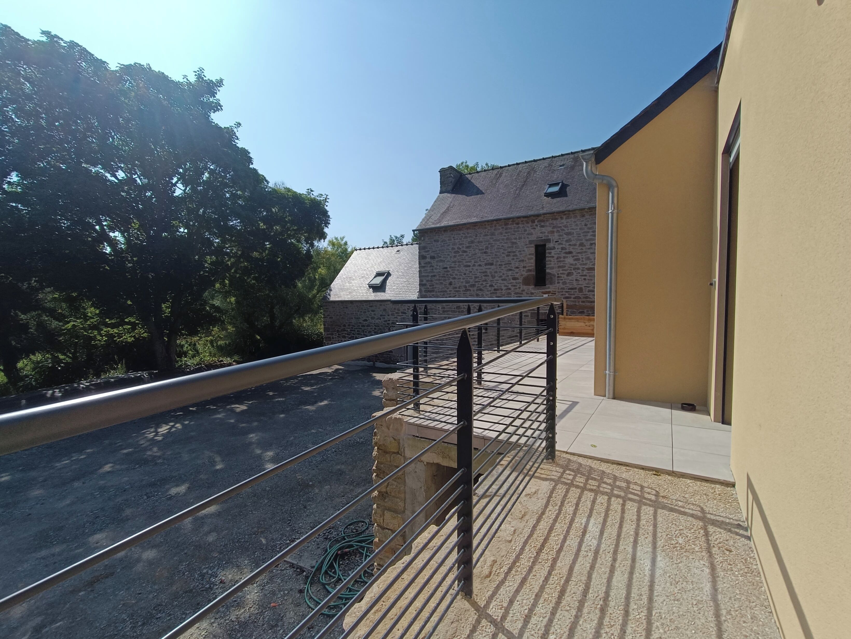 A balcony with a metal railing overlooking a gravel driveway and a stone building with a sloped roof.