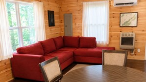 A cozy cabin interior with a red sectional sofa, a round wooden dining table, and a small kitchen area with an air conditioner.