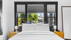 Una habitación de hotel moderna con una cama grande, dos mesitas de noche y vistas a una zona residencial a través de amplios ventanales.
