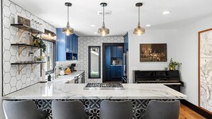 Una cocina moderna con una isla de mármol, armarios azules, iluminación colgante y un piano.