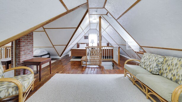 A loft bedroom with a sloped ceiling, wooden beams, a brick wall, a bed, a sofa, and a small table. - Pondside Retreat Near Narrowsburg & The Delaware River (Beach Lake)