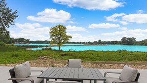 Zona de estar al aire libre con una mesa y sillas, con vista a un cuerpo de agua turquesa y un paisaje de césped.