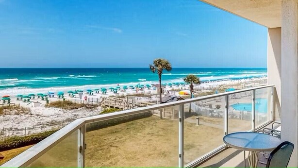 Un balcón con vistas a una playa, una piscina y un cielo azul despejado.