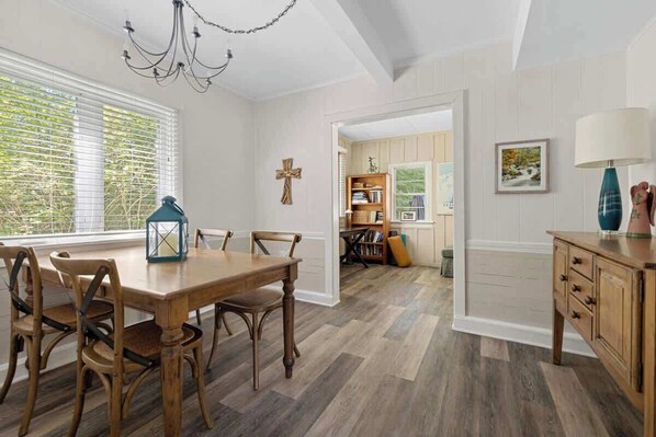 A dining area with a wooden table and chairs, a chandelier, and a view through a window with blinds.