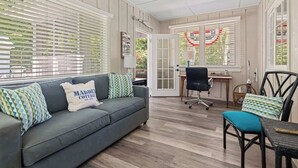 Een kamer met een grijze bank, een blauwgroene fauteuil, een bureau met stoel en een raam met zonwering.