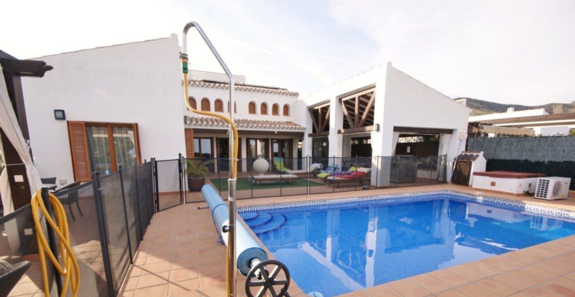 A swimming pool with a clear view of the surrounding area, including a building with a tiled roof and a fenced patio area.