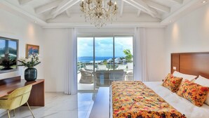 Ein Hotelzimmer mit einem großen Bett, einem Schreibtisch, einem Sessel, einer Kandelaber und einem Balkon mit Meerblick.