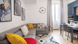 Una sala de estar moderna con un sofá seccional gris, un comedor con una mesa dispuesta para dos personas, y un reloj de pared grande.