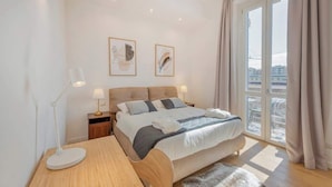 Un dormitorio con cama, mesitas de noche, un escritorio y un amplio ventanal con cortinas.