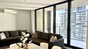 A modern living room with a sectional sofa, a dining table with wine glasses, and large windows offering a city view. - High Floor 2BR2Bath in Prime CBD (Melbourne)