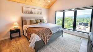 A bedroom with a large bed, a nightstand, a window with a view of a forest and buildings, and a wooden ceiling. - Gorgeous 4BD Lodge w/ POOL! Hot Tub + Theater (Sevierville)