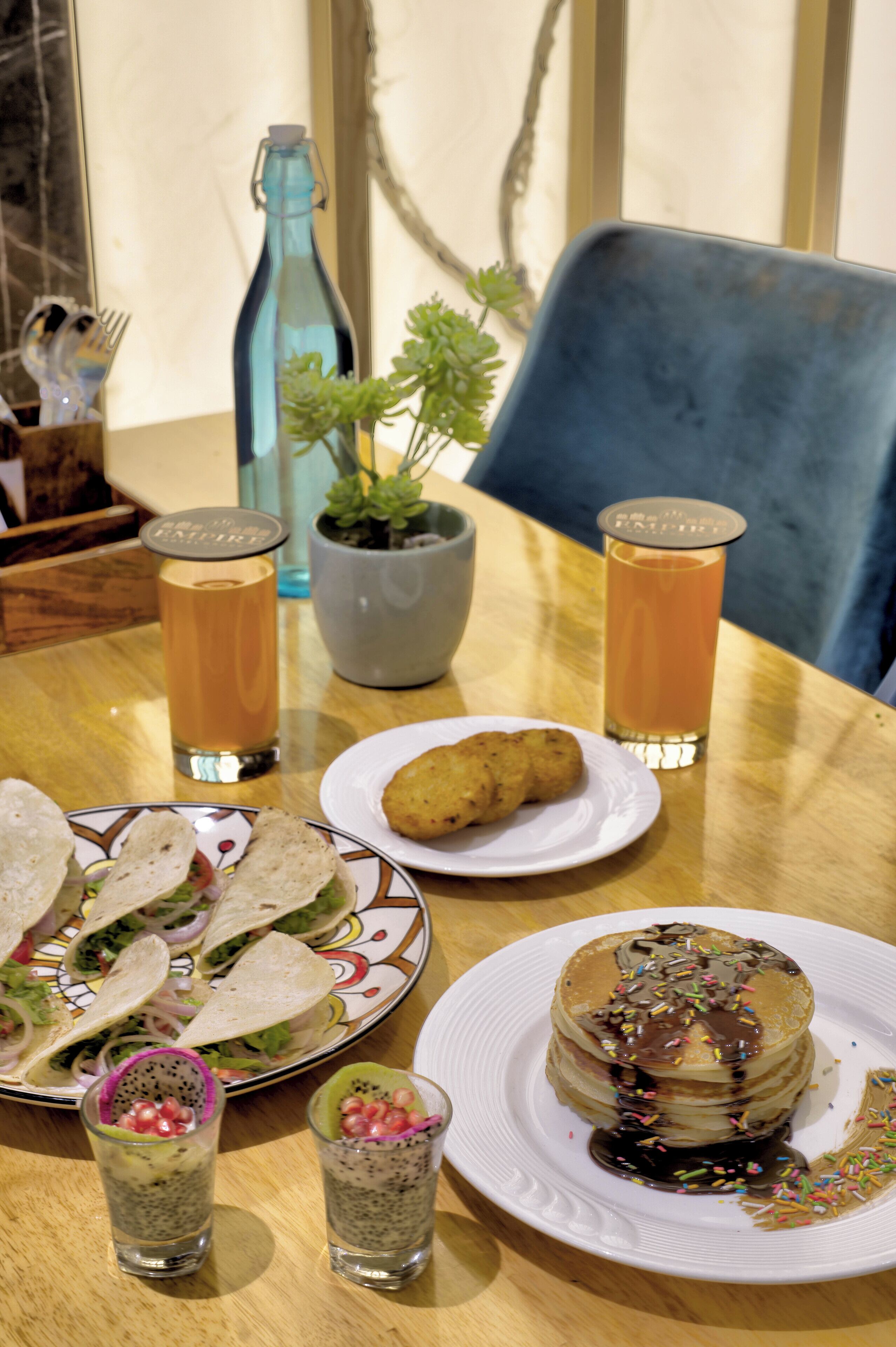 A dining table with a plate of tacos, a plate of pancakes with chocolate syrup, two glasses of orange juice, a bottle of water, and a potted plant.