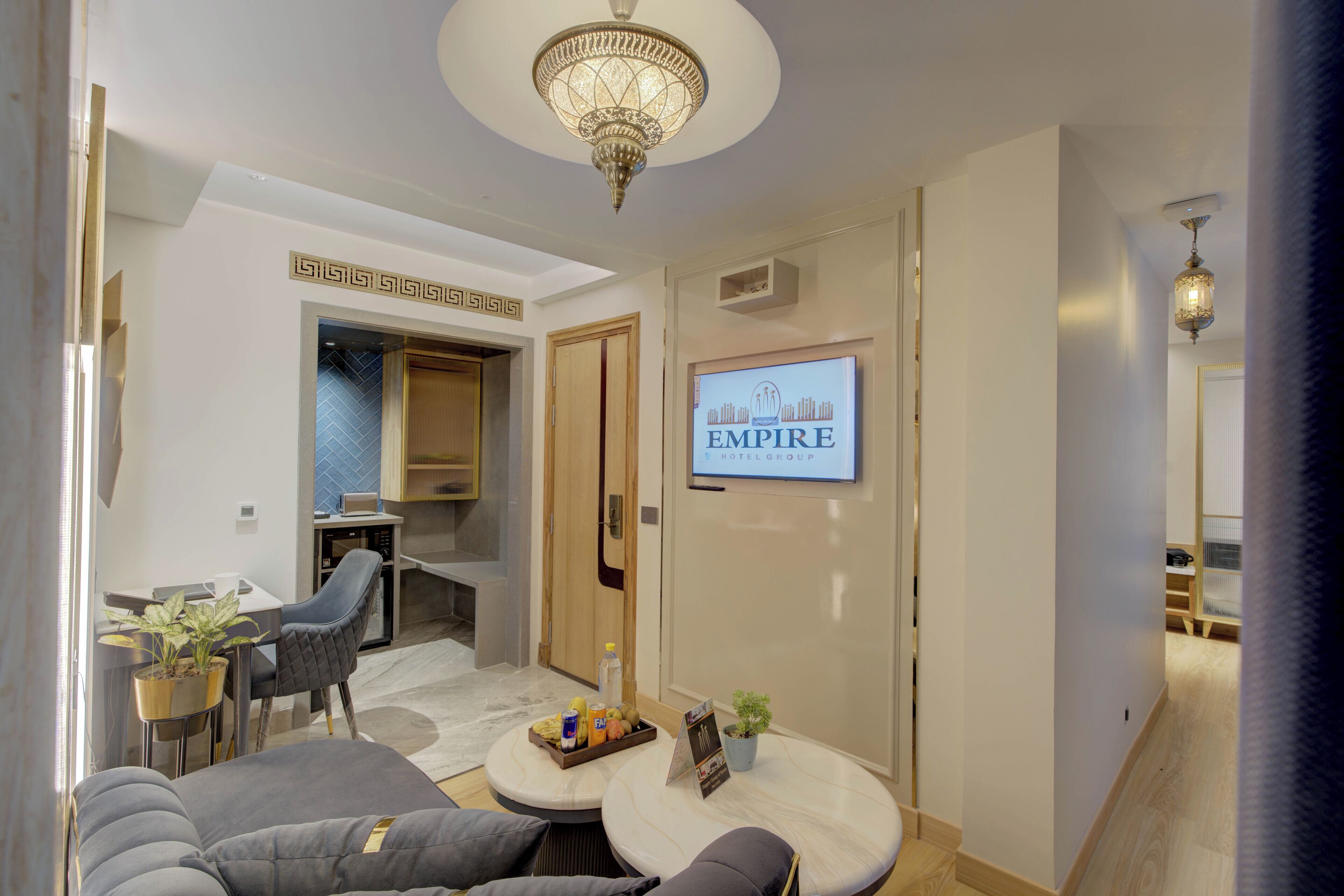 A hotel lobby with a modern design, featuring a seating area with sofas and a coffee table, a television displaying the hotel name, and a chandelier.