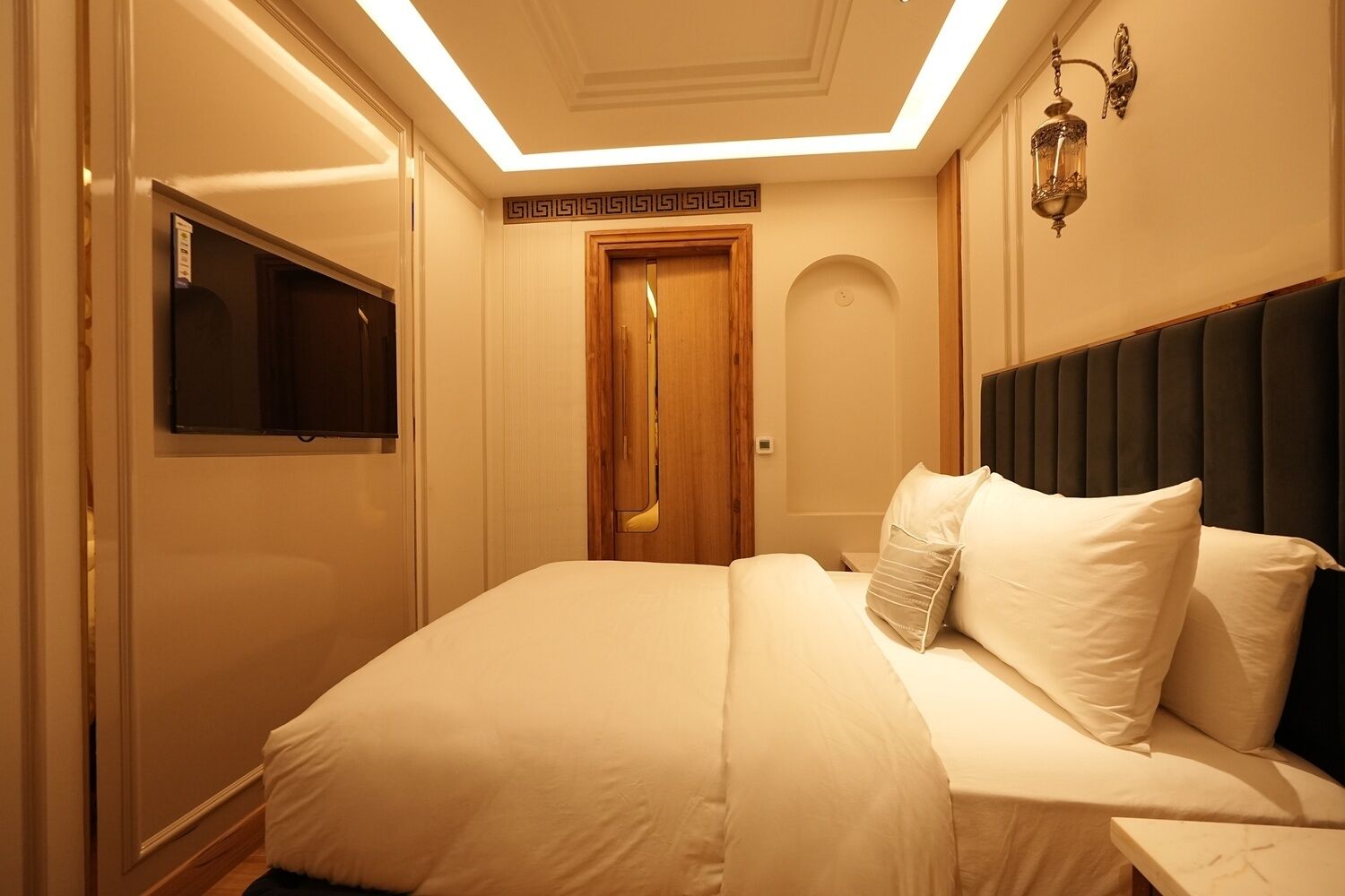 A hotel room with a bed, a television, a wooden door, and a wall-mounted light fixture.