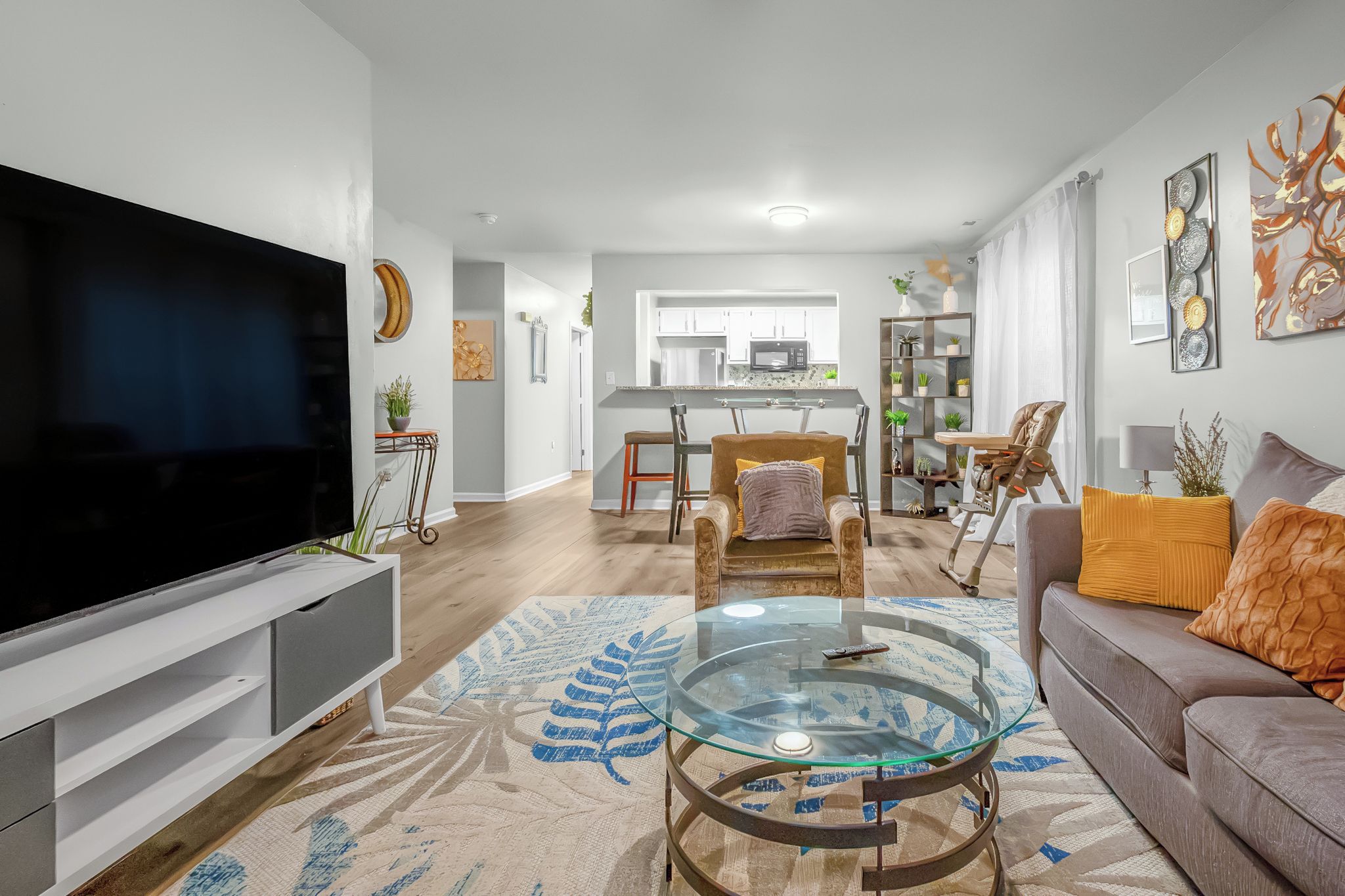 A modern living room with a flat-screen TV, a glass coffee table, a sofa with cushions, and a dining area with chairs.