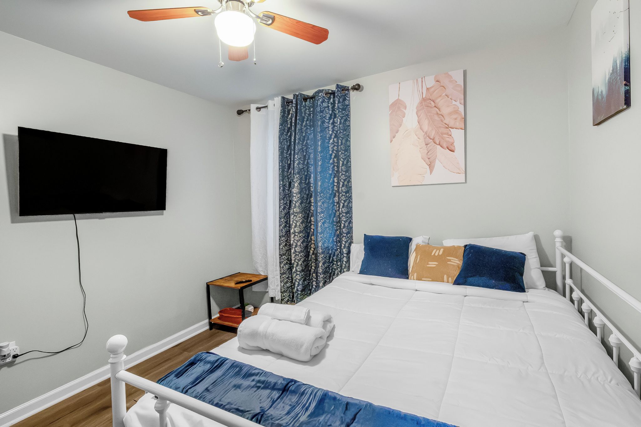 Une chambre à coucher avec un lit, un téléviseur fixé au mur, un ventilateur de plafond et une fenêtre avec des rideaux.
