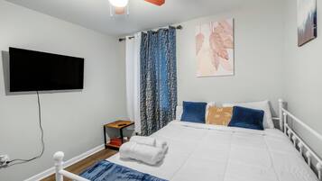 Une chambre à coucher avec un lit, un téléviseur fixé au mur, un ventilateur de plafond et une fenêtre avec des rideaux.