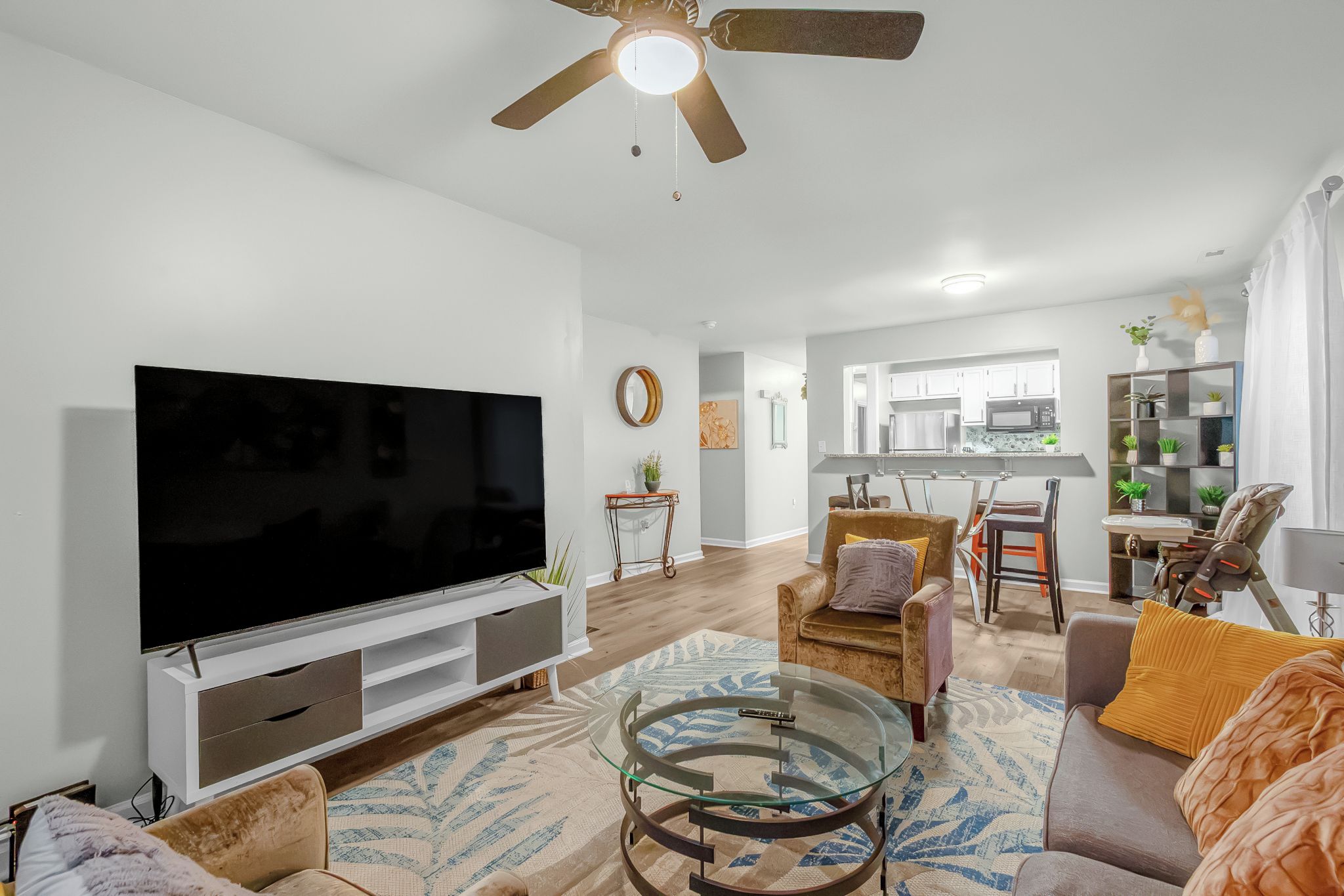 A modern living room with a flat-screen TV, a glass coffee table, and a ceiling fan.