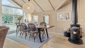 Una habitación acogedora con una mesa de comedor, sillas, una lámpara colgante, chimenea y una ventana que ofrece vistas al verdor.