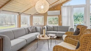 Una habitación acogedora con un sofá gris, sillas de mimbre, una mesa redonda y ventanas amplias.