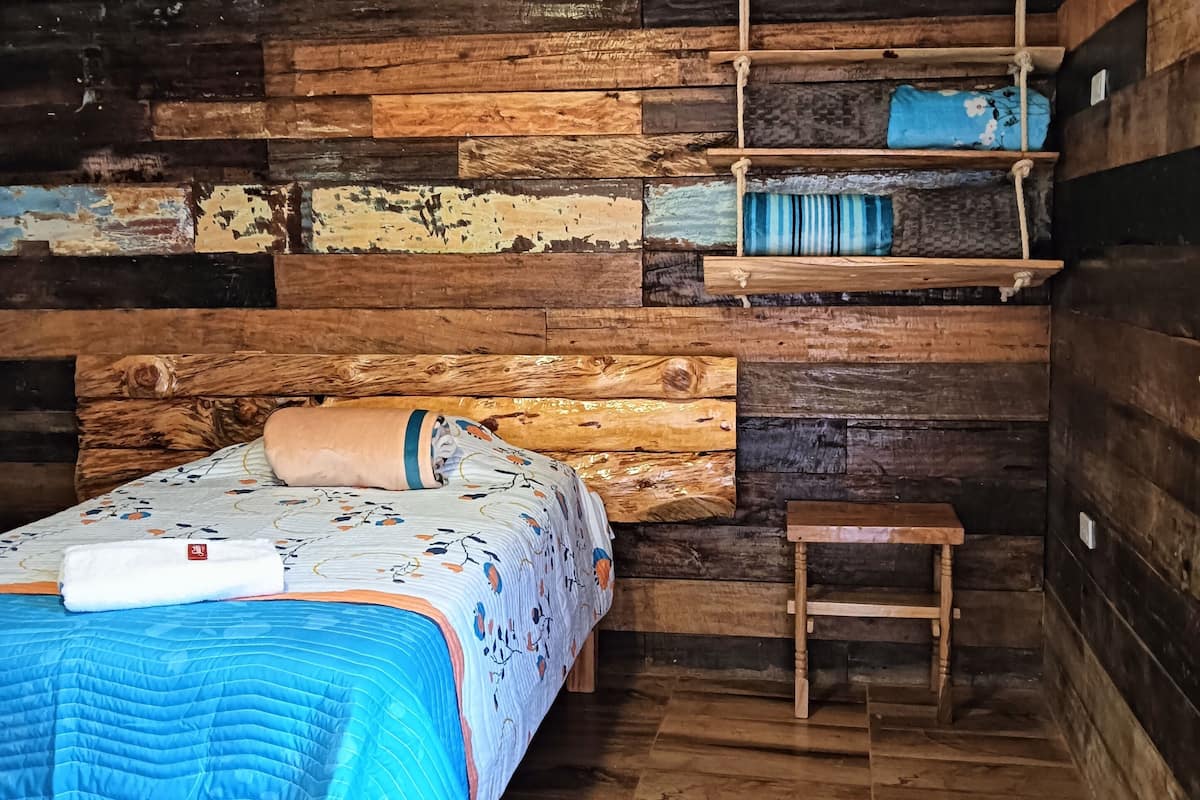 A rustic cabin-style room with a wooden bed, a small wooden bench, and a shelf with pillows.