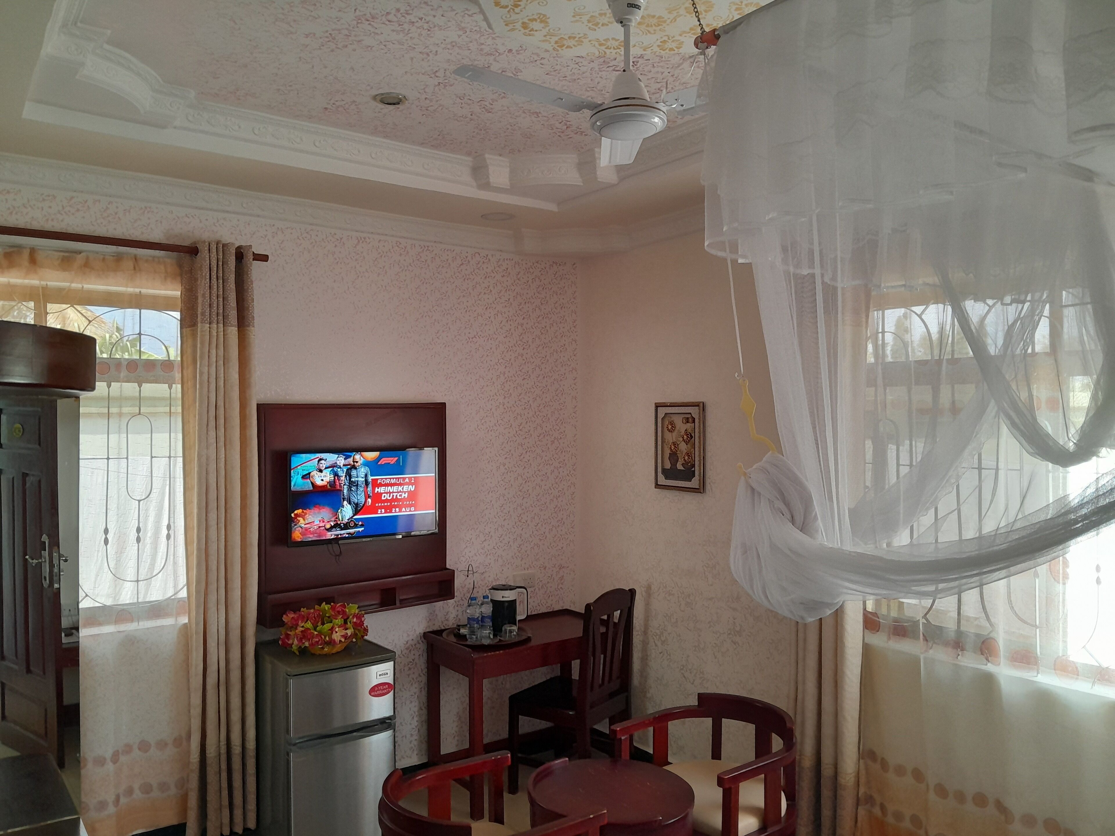 Una habitación con televisión mostrando un evento de Fórmula 1, ventilador de techo, una mesa con sillas y ventana con cortinas.