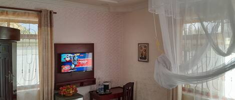 Una habitación con televisión mostrando un evento de Fórmula 1, ventilador de techo, una mesa con sillas y ventana con cortinas.