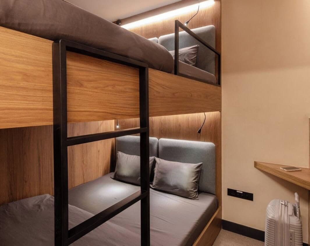 A bunk bed with a wooden frame and grey bedding, a small bedside table with a lamp, and a suitcase on a shelf.